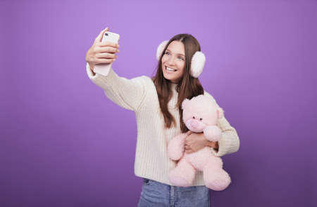 A girl with dark hair in a light sweater and furry pink ears takes a selfie and cuddles with a pink toy bear. Photo on a lilac background. The atmosphere of fun. Teenagerの写真素材