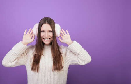 Bright positive girl in a knitted sweater and fur headphones on her head holds her hands behind her head. He looks straight and smiles broadly. Playful mood. Winter clothes. Photo on a background.の写真素材