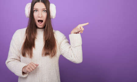 A surprised girl with dark hair, with bright emotions, in a knitted sweater and fur headphones shows a finger to the side. He looks straight, opening his mouth. Photo on a purple background.の写真素材