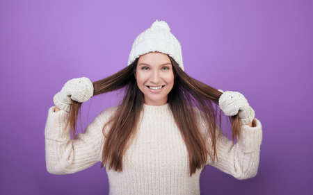 Smiling girl in a winter white knitted hat. A young girl in a good mood in a warm white sweater and white mittens. Good New Year spirit. Photo on a purple background.の写真素材