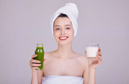 Cute fresh girl in a towel on her head, after a shower, half-naked holds milk in her hands and a green smoothie to choose from. With a wide smile, looking straight. Body cleansing. Detox. Diet.の写真素材