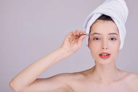Beautiful half-naked girl wrapped in a white towel after a shower plucking eyebrows with tweezers. Holds tweezers with his right hand near the eyebrow. Style and fashion. Hair Removal, Laser.の写真素材