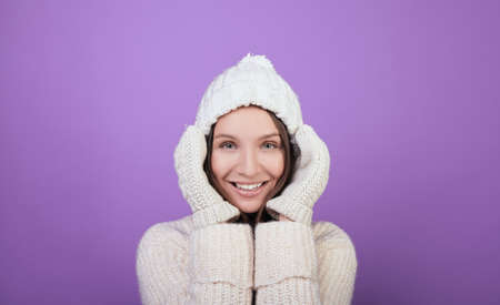 Charming girl with green eyes on a winter white knitted hat. Photo of a young girl in a good mood in a warm white sweater and white mittens. Christmas mood. Photo on a purple background.の写真素材
