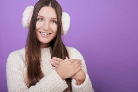 Cute girl with green eyes, dark hair, in a knitted sweater and fur headphones folded her arms over her chest. He looks at the camera and smiles. Photo on purple background. The atmosphere of comfort.の写真素材