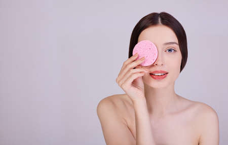 A girl with dark hair sensually holds a pink sponge near her face. Portrait of a young girl on a gray background. Youth and skin care concept.の写真素材