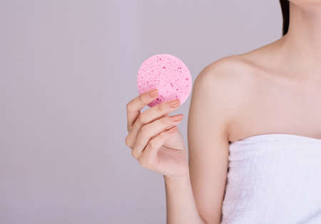 Attractive woman with dark tied hair holds a makeup sponge on her face, takes care of her face, removes pores, poses half naked, smiles gently against a light background.の写真素材
