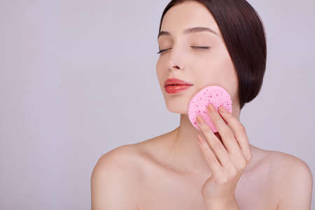 Attractive woman with dark tied hair holds a makeup sponge on her face, takes care of her face, removes pores, poses half naked. Personal care.の写真素材