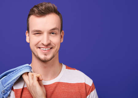 Radiant smiling beauty man in striped t-shirt stands in the background, looks straight and winks with one eye. In one arm behind a guy holds a denim shirt. Close up, copy space, large frame.の写真素材