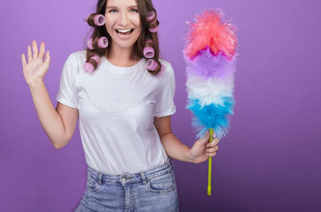 A bright girl with dark hair and green eyes in curlers holds a dust brush in her hand and laughs. Fun while cleaning. Housework. Cleaning. A maid or housewife is cleaned.の写真素材