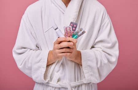Man in a white coat in a blue scarf with an enema in one hand and pills and a inhaler in the second. Ill, illness, temperature, obstruction. Medicine, drugs. Closeup.の写真素材