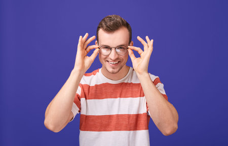 Young cheerful student, employee, guy in striped T-shirt with round glasses, holds on to glasses...