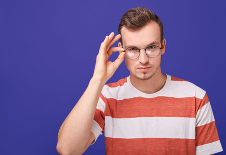 A young charismatic guy in a striped T-shirt bowed his head forward and holds round glasses with his...