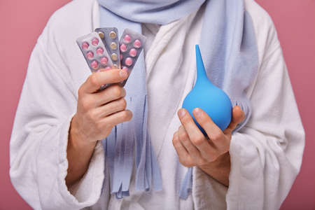 Illness man stands on pink background, holds an enema in right hand and in the right pills. Recovery, treatment, procedures, poor health. White bathrobe, blue scarf.の写真素材