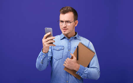 Pensive businessman looking attentively on a gray mobile phone. A man in glasses, a blue denim shirt with a notebook and a pen in his hand. Student, teacher, entrant, employee.の写真素材