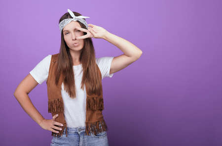 A cute girl with green eyes in a white T-shirt, brown vest and headband shows with his left hand the peace sign and makes a kiss. Peace for peace. Style 90. Retro style. Children of flowers.の写真素材