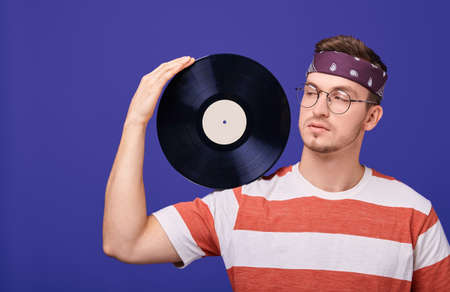 A nice handsome guy with round glasses, a striped T-shirt, a bandana on a head and holding one black round music record with one hand. Playfully looks away. Retro style. Style 90. Photo isolate.の写真素材