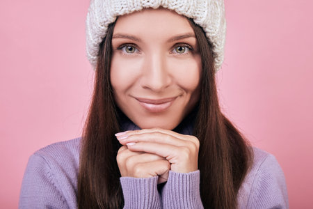 Cute girl with dark hair in white knitted hat clasped her hands under her chin. She looks down and...