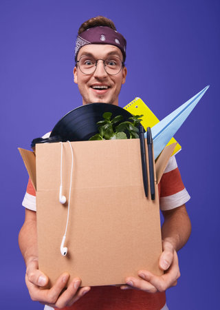 Joyful young guy in round glasses and a brown bandana on his head holds in his hands a box with...