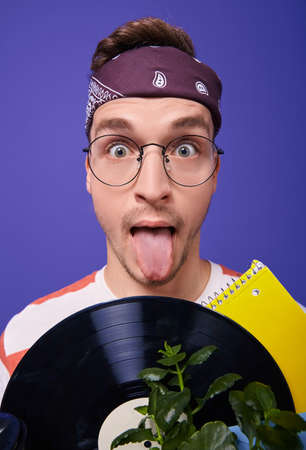 A cheeky and bright guy in round glasses and a brown bandana on his head holds in his hands a box with things, shows his tongue. Paper plane, vinyl record. Young designer, musician.の写真素材