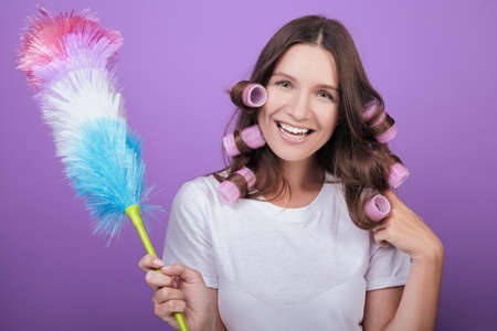 A cheerful housewife with a wide snow-white smile in pink curlers and a white T-shirt brushes the dust with a pipidaster. Posing on a purple background. Cleaning and cleanliness.の写真素材