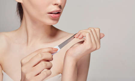 Young beautiful smiling girl sits on a background holds a nail file in her right hand and sharpens her nails on her left hand with enthusiasm and looking on her nails. Cosmetology.の写真素材