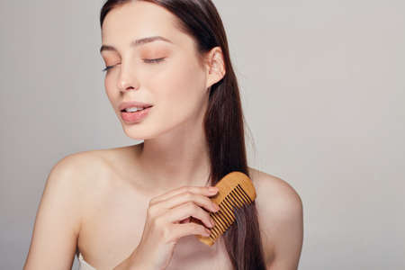 A tender calm pretty girl with closed eyes combs her hair with a light brown comb on the gray background. Lady looks away at left side. Spa, body care. Copy space.の写真素材