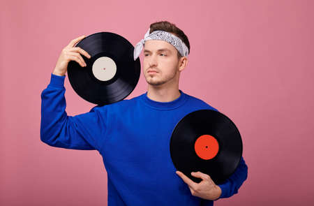 Close-up. A cool stylish guy in a bandana in a dark blue jacket stands on a pink background with a black vinyl record in his hand. Youth, style, joy, playful mood. Retro style.の写真素材