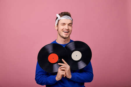 Happy smiling stylish guy in bandana in a dark blue jacket stands on a pink background with two black vinyl records in his hands. Youth, style, joy, playful mood. Retro style. Close-up. Focus on vinilの写真素材