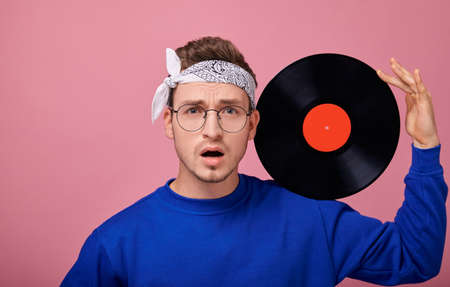 Focus on vinil. Close-up. A cool stylish guy in a bandana in a dark blue jacket stands on a pink background with a black vinyl record in his hand. Youth, young, style, confidence, joy, surprised.の写真素材