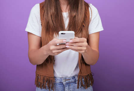 A girl in a white T-shirt, a brown vest, with dark hair, is holding a phone in a white case with both hands. SMS correspondence. Close-up. Social networks. Photo on a purple background.の写真素材