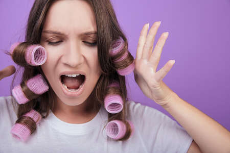 Woman screaming and shocked. The lady raises her hands with great fear. She has curlers in her hair. She is in a white t-shirt. Photo on a purple background.の写真素材