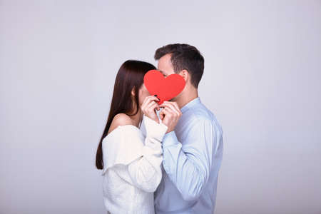 Beautiful couple of lovers stands on a white background covering their faces with a red heart. A brown-haired guy in a blue shirt and a dark-haired girl in a snowy white sweater. Valentine's dayの写真素材