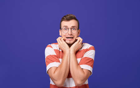 Handsome young guy in a striped t-shirt posing on a blue background. Frightened, holds his hands near his face, showing fear. Photo isolate on background.の写真素材