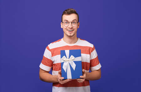 A young guy with blond hair in glasses and a striped T-shirt is holding a gift box in his hands. Blue gift with a white ribbon. Photo isolate on a blue background.の写真素材