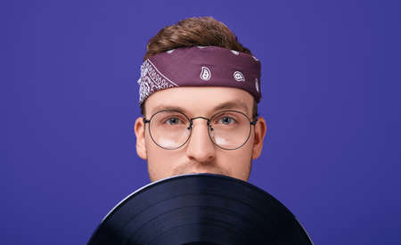 A confident and calm guy with round glasses, a striped T-shirt, a bandana on a head covered his face with a musical record. Looks straight. Retro style. Style 90. Photo isolate. Close-up.の写真素材
