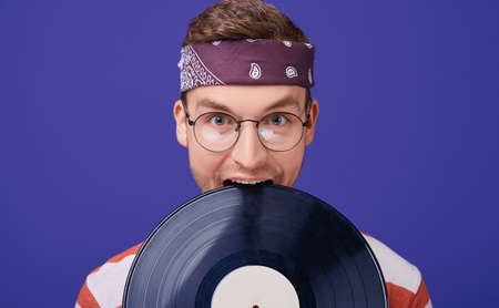A handsome guy in round glasses, a striped T-shirt, a bandana on a gag jokingly bites a music record. The emotion of fun and joy. Retro style. Style 90. Photo isolate. Close-up.の写真素材