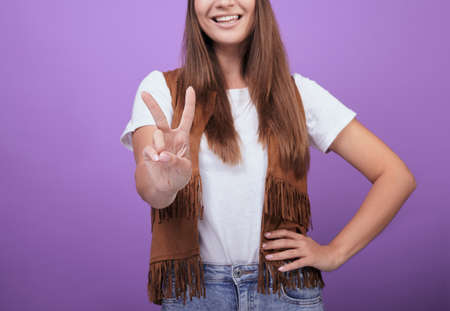 Smiling woman in a brown vest and a white T-shirt with brown beautiful hair shows her fingers a peace sign close-up. Photo on a purple background. The concept of peace and pacifism.の写真素材
