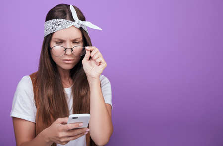 A young student in a white T-shirt and round glasses holds a phone in her left hand, reads SMS. Holds a temple of glasses. Vivid emotions of surprise and doubt. Photo on a purple background.の写真素材