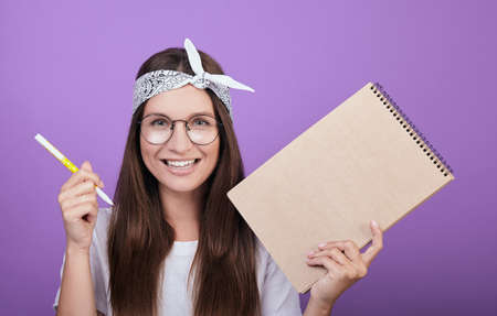 A friendly girl with dark hair with a smile, in a white T-shirt and round glasses, holds a pen and craft notebook in her hands. Retro style. Photo on a purple background.の写真素材
