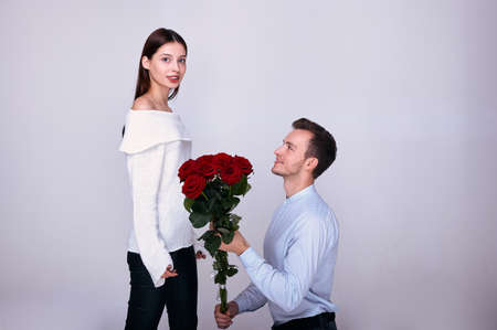 Elegant man in a blue shirt with a bouquet of red roses kneeled and gives his beautiful girl in a white sweater red roses. The girl smiles and looks at the camera. Valentine's Day. Marriage proposal.の写真素材