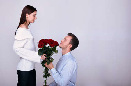A handsome guy in a blue shirt with a bouquet of red roses knelt down and makes a marriage proposal to his beautiful girlfriend in a white sweater. Lovers look at each other. Valentine's Day.の写真素材