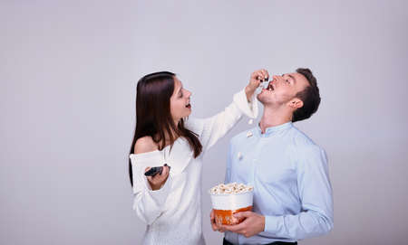 Beautiful young couple in love while watching movie at the home theater frolic. The girl holds the remote control from the TV and spills popcorn into the guy's mouth, and guy holds a basket of popcornの写真素材