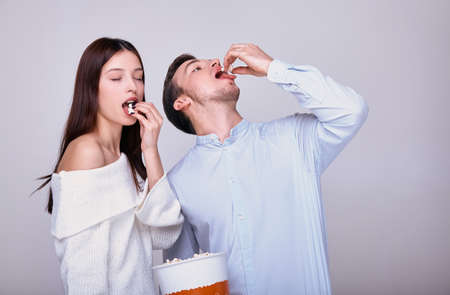 Young couple on a gray background eating popcorn. The girl closed her eyes in satisfaction, and the guy throws popcorn directly into her mouth. Family weekend. Cinema and theater. Time together.の写真素材
