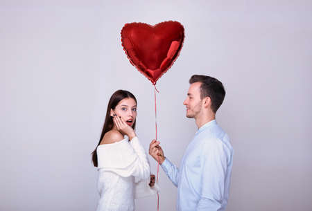 A young blond in a blue shirt gives the girl a red heart-shaped balloon, the girl takes a gift and is very surprised, holds her hand near her mouth. Valentine's Day. Birthday. Surprise gift.の写真素材