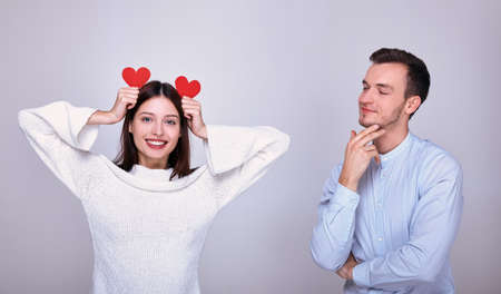 A sexy girl with a sweet smile, dark hair and blond eyes, in a white knitted sweater holds two red hearts in her hands. A charming man stands next to a beautiful lady and looks at her with a smile.の写真素材
