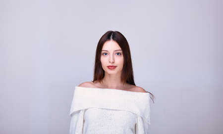 A young girl with dark hair, puffy lips, blond eyes, with bare shoulders in a knitted white sweater calmly stares straight ahead. Photo on a light background. IThe emotion of confidence and sexuality.の写真素材
