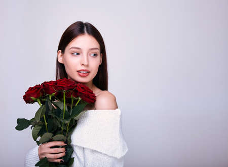 Charming tender girl with dark hair, fair skin, in a white knitted sweater with bare shoulders, holds red roses in her hands and looks thoughtfully to the side. Photo on a gray background.の写真素材