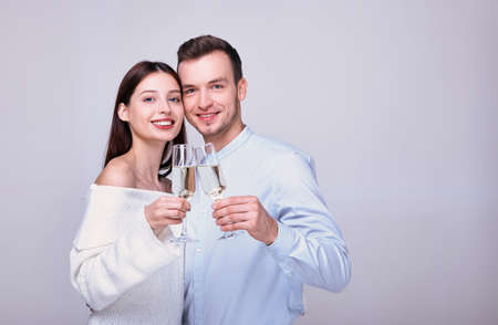 A young guy in a blue shirt smiles happily and raises a glass of champagne. Nearby stands an elegant lady with a snow-white smile and also holds a glass in hands. A couple celebrates Valentine's Day.の写真素材