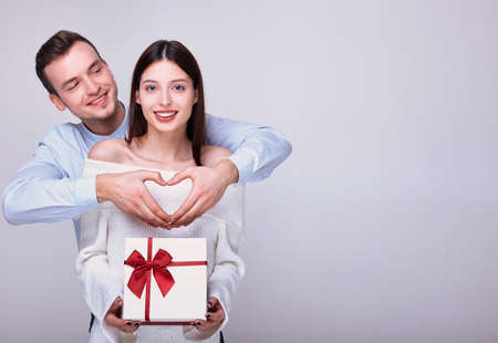 A cheerful young man in blue shirt is standing behind cute attractive smiling girl in snow-white sweater with gift in her hands for Valentine's Day. Guy shows heart sign with his own hands. Copy spaceの写真素材