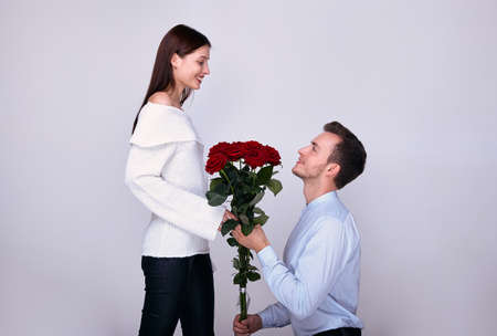 A young guy in a blue shirt knelt and gives his beautiful girlfriend in a white sweater a bouquet of red roses. Lovers look at each other. Valentine's Day. Marriage proposal.の写真素材